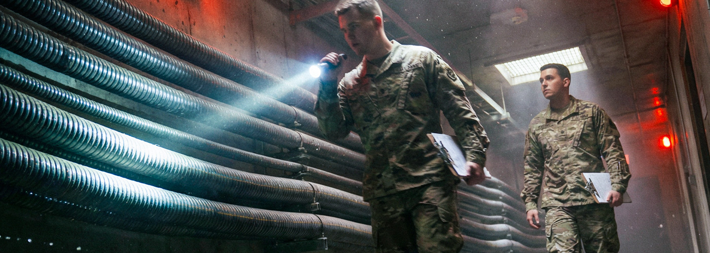 Two soldiers manually inspect fiber optic conduit along a corridor.